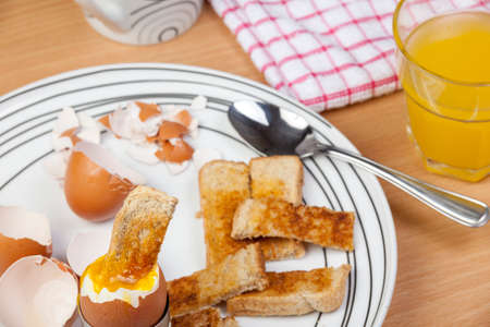 Dipped runny egg soldier on a plate with broken egg shells and a  glass of orange at the side