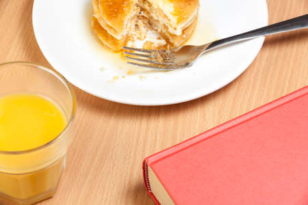 Stack of part eaten pancakes covered in syrup on a breakfast table with a book and a glass of orange juiceの写真素材