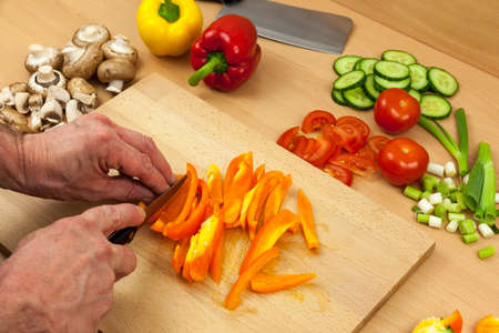 Close up shot of a chefs hands slicing a cleaned orange bell pepper on a wooden chopping board aside a selection of chopped mixed vegetablesの写真素材