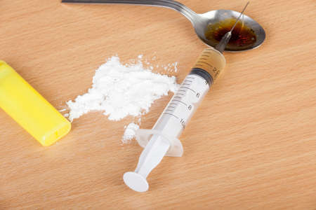 Loaded heroin syringe laying across a burnt spoon on a table with a pile of heroin and a lighter