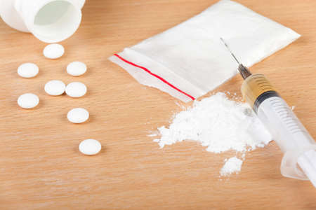 Loaded syringe laying on a bag of heroin and some spilled pills on a table
