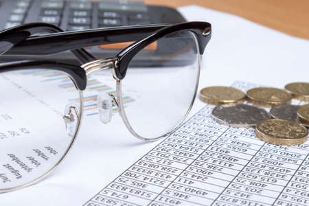 Close up shot of a pair of black rimmed spectacles laying on an account's desk with some loose change and a calculatorの写真素材
