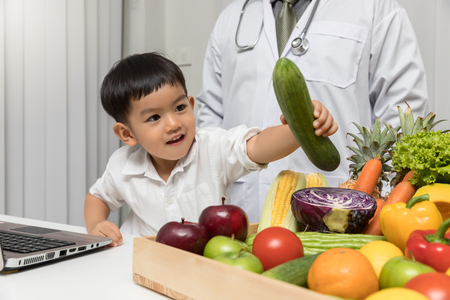 Healthy and nutrition concept. Kid learning about nutrition with doctor to choose eating fresh fruits and vegetables. Young asian boy holding cucumber.のeditorial素材