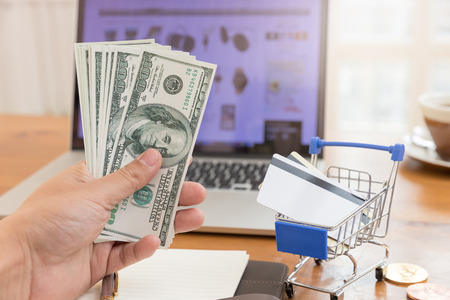 E-commerce concept, hand holding money and credit card in shopping cart with laptop open shopping website background.の写真素材