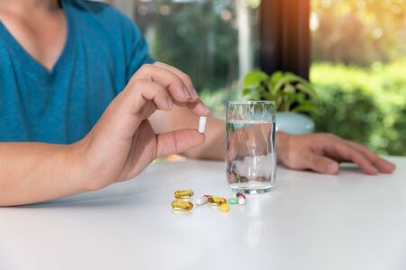 Young man holding assorted pharmaceutical medicine pills in one hand and glass of water in other oneの写真素材