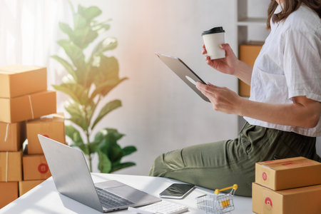 Concept of parcel delivery and selling online,Retailer Using computer tablet to check sales for the day on an online store while holding coffee cup with parcel box that prepare to send to customerの写真素材