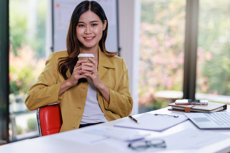 Concept of business office woman working,Businesswoman smile while working about her invesment plan with analyzing document and business investment graph data and holding coffee cup in workstationの写真素材