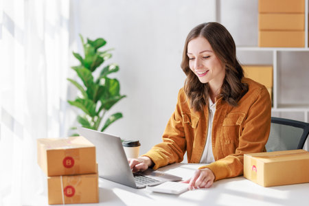 Concept of parcel delivery and selling online, Female seller or retailer using laptop to check sales of the day on online store to be packed into the parcel and ready to deliver to the customerの写真素材