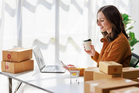 Concept of parcel delivery and selling online, Female seller or retailer using laptop to check sales of the day on online store to be packed into the parcel and ready to deliver to the customerの写真素材