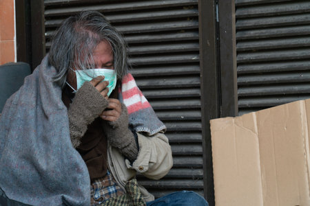 Homeless man with face mask sitting in front of a closed doorの写真素材