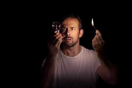 Worried man looking at a light bulb with a match in the dark. Concept of blackout. Selective focus.の写真素材