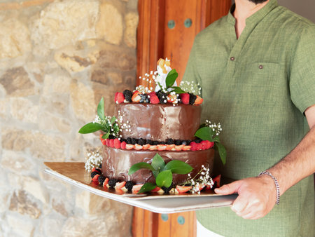 A person holds a two-tier chocolate cake decorated with fresh berries and leaves.