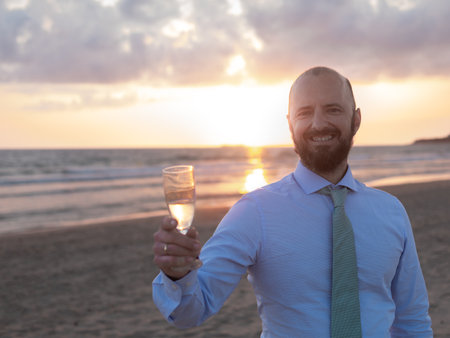 A formally dressed man holds a champagne glass on a beach, with the setting sun casting a warm glow.の写真素材