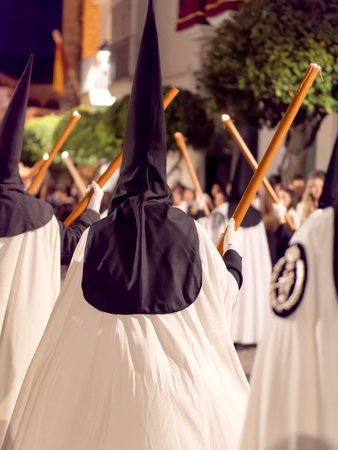 Nazarenes in white tunics and black hoods walking in a night procession with candles.