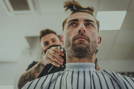 a young man with tattooed arms cuts a man's hair in a barbershopの写真素材
