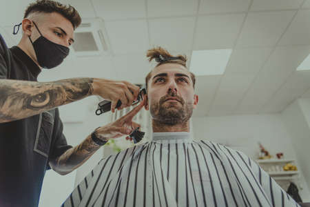 a young man with tattooed arms cuts a man's hair in a barbershopの写真素材
