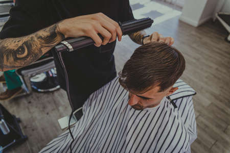 a young man with tattooed arms cuts a man's hair in a barbershopの写真素材