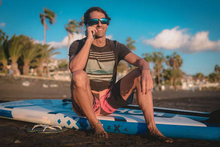 a young man uses his mobile phone sitting on his paddle board on the beach