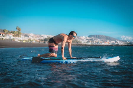 a young sportsman man practices paddle surfing on the beach under a blue skyの写真素材