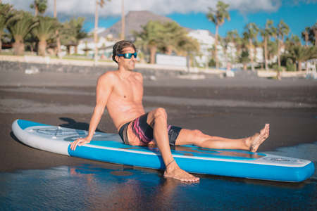 a young sportsman man rests after paddle surfing on the beach under a blue sky