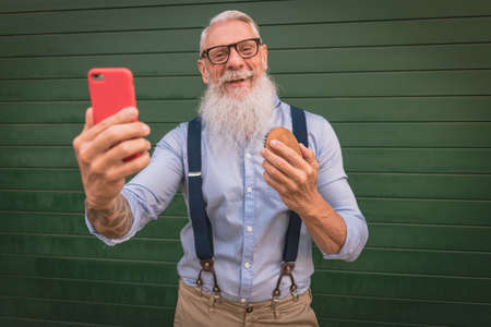 an older man in hipster clothes and glasses and a long white beard comb his beard and take a photo focus on headの写真素材