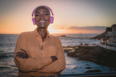 a black adult woman is listening to happy music, she sees the sunset and the sun rays with the sea...