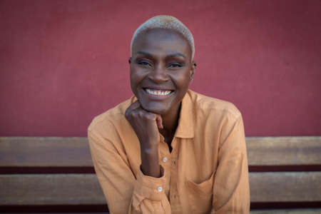 A black adult woman is sitting on a wooden bench and a red wall is in the background. She is relaxed. Focus on the faceの写真素材