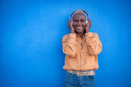 an adult black woman poses with headphones on a blue wall and smilesの写真素材