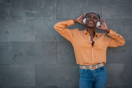 an adult black woman poses with headphones on a gray wall and smiles she is happyの写真素材