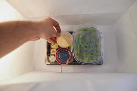 From above anonymous hand picking up a sauce from a paper bag of sushi