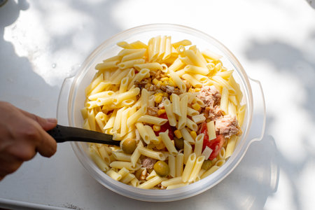 From above anonymous person taking portion of tasty pasta with tuna, vegetables and olives from plastic bowl on sunny dayの写真素材