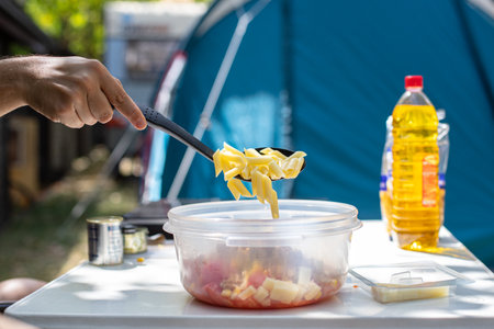Anonymous male adding penne pasta into bowl while making pasta salad on campsite in summerの写真素材