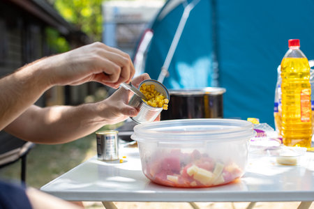 faceless male sitting at table adding canned corn into plastic bowl with salad cooking lunch on campsiteの写真素材