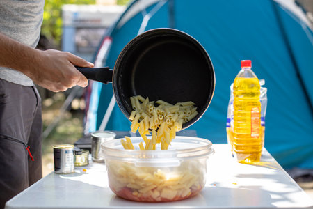 Unrecognizable man putting fresh penne pasta from saucepan into plastic bowl during lunch preparation on campsiteの写真素材