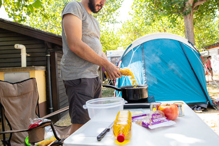 Bearded man in shorts standing putting pasta in a saucepan preparing food in a camp on a summer dayの写真素材