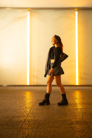Full body side view of stylish female in black outfit and leather jacket walking along illuminated corridorの写真素材