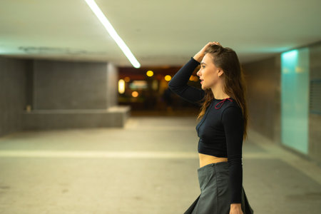 Side view of young female with long dark hair in black top and skirt standing in parking building and touching headの写真素材