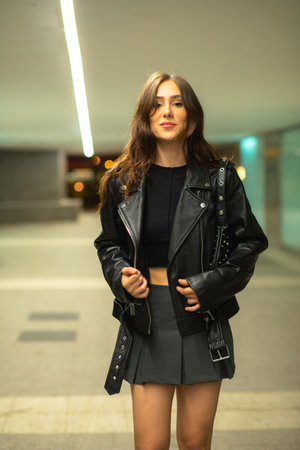 Positive young woman with long wavy in black top skirt and leather jacket standing in parking building and looking at cameraの写真素材