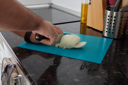 Anonymous person cutting an onion on a blue cutting board on a black countertop in a lighted kitchenの写真素材