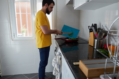 Side view of a young bearded man putting chopped vegetables in a frying pan in a brightly lit kitchenの写真素材