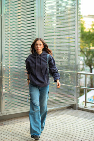 Full body of confident young woman in trendy jeans and hoodie walking on wooden footbridge against cityscapeの写真素材