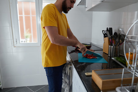 Side view of a young bearded man cutting a red bell pepper in the kitchenの写真素材