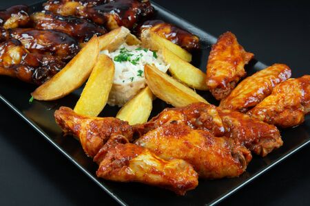 Close up of chicken wings with barbecue sauce and hot sauce, accompanied by French fries and German salad, in a rectangular black plate, isolated on a black background. Concept of tasty and popular food.の写真素材
