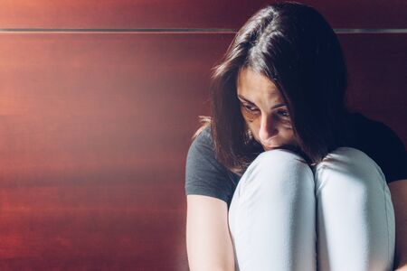 Young girl, victim of domestic violence, sitting, with her head on her knees, looking at the floor,blank stare. Bruises and stitches under her eyes. Soft side light. Copy space. Concept of gender violenceの写真素材