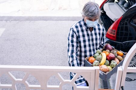 Man with gray hair, wearing an antibacterial mask and sanitary gloves, holds a box of fruits and...