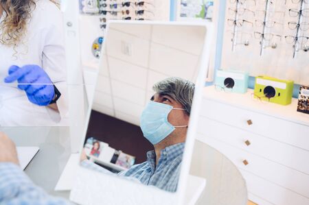 Gray-haired man with a face mask reflected in a mirror while talking to the especialist doctor in an...
