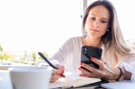 Beautiful young girl, successful entrepreneur looking at her mobile phone and writing in her notebook. Copy space. Business and young entrepreneurs conceptの写真素材