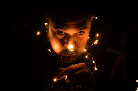 Portrait of enigmatic young man holding Christmas lights over his face. Isolated on a black background. Copy space. Concept of enigma and mysteryの写真素材