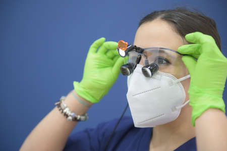 Attractive young female dentist wearing sanitary mask putting on binocular medical magnifier. Blue background.Copy space.Women workers and technical equipment conceptの写真素材
