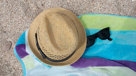 Yellow straw hat shade towel of blue and green colors on the white sand and next to sunglasses